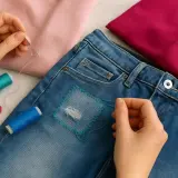 Hände flicken mit Nadel und Faden eine Jeans. Garnrollen und Knöpfe liegen bereit.