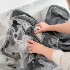 Hände waschen ein graues Schurwollkleidungsstück in schaumigem Wasser. Sanft wird die Wolle gereinigt, um sie frisch zu halten.