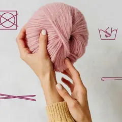 Hände halten rosafarbene Wolle. Symbole zeigen Handwäsche, kein Trocknen und Stricknadeln/Häkelnadel.