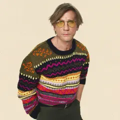 Mann mit gelber Brille trägt bunten wollpullover. Er fragt sich, wie er diesen am besten waschen kann, um die Farben zu erhalten.