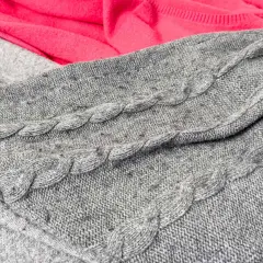 Grauer Strickpullover mit Zopfmuster und ein pinker Stoff. Kleine Knötchen zeigen, wie man Pilling vermeiden kann.
