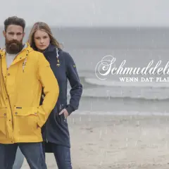Mann und Frau am Strand im Regen, er trägt eine gelbe Schmuddelwedda Jacke, sie eine blaue. Perfekt für jedes Wetter!