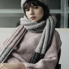 Junges Mädchen mit einem gestrickten Schal aus Wolle und passender Mütze. Der dicke, graue Schal ist um den Hals geschlungen und reicht bis zum rosafarbenen Pullover.