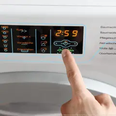 Finger drückt auf Start/Stop an Waschmaschine. Display zeigt 2:59. Wähle das richtige Programm für Wolle, achte auf die richtige Temperatur.