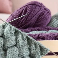 Stricken mit dicker Wolle: Ein Strickprojekt mit Zopfmuster in Grau und Lila, Nadeln stecken im Gestrick.