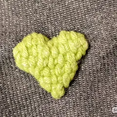 Ein gehäkeltes Herz in leuchtendem Grün bedeckt ein kleines Loch im Stoff. Ein unsichtbarer Flicken, der Liebe zeigt.