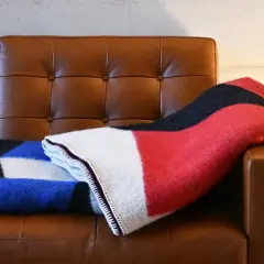Zwei Decken auf braunem Ledersofa: eine mit geometrischem Muster in Blau, Schwarz und Weiß, die andere in Rot, Schwarz und Weiß. Der Unterschied zwischen Wolle und Baumwolle ist spürbar.