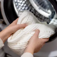 Hände legen einen weißen Strickpullover in die Waschmaschine, um eingelaufene Kleidung zu retten.