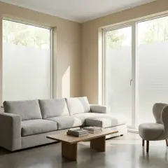 Helles Wohnzimmer mit grauem Sofa, Couchtisch und Sessel. Die Fenster sind mit Milchglasfolie beklebt, die das Licht sanft filtert. Die Folie ist so gestaltet, dass sie wie feine Gardinen färben wirkt.