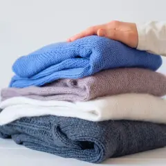 Ein Stapel gestrickter Pullover in verschiedenen Farben, von dunkelblau bis weiß. Eine Hand streicht sanft über den obersten Pullover, als ob er per Hand gewaschen wurde.
