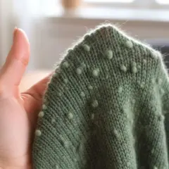 Grüner Strickpullover mit Noppenmuster, gehalten von einer Hand. Die feine Merinowolle fusselt leicht am Rand.