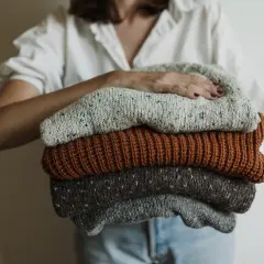 Eine Person hält einen Stapel gestrickter Pullover. Die Pullover sind in Grau-, Braun- und Beigetönen gehalten. Sie sind bereit, um **Wolle bei 40 Grad waschen** zu können.
