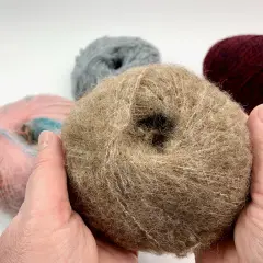 Hände halten ein Knäuel beiger Wolle, bereit, einen Schal zu stricken. Weitere Wollknäuel in Rosa, Grau und Bordeauxrot liegen im Hintergrund.