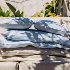 Stapel Kissen in Blau und Beige. Der feine Unterschied zwischen Leinen und Baumwolle ist spürbar. Schatten von Blättern fallen darauf.