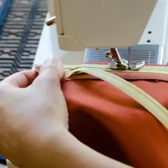 Hände nähen einen Reißverschluss mit einer Nähmaschine an ein orangefarbenes Kleidungsstück. Reißverschluss reparieren leicht gemacht.
