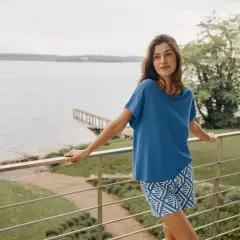 Frau auf Balkon mit Blick aufs Wasser, trägt blaues Oberteil und gemusterte Shorts aus Modal Stoff. Waschen ist einfach.