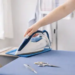 Eine Hand hält ein Bügeleisen über einem Bügelbrett. Man fragt sich, ob Leinen beim Waschen eingeht, aber hier wird es glatt gebügelt.