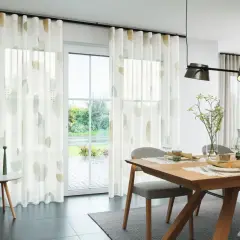 Ein Esszimmer mit Holztisch und Stühlen. Ein Vorhang nur auf einer Fensterseite mit abstrakten Mustern.