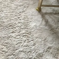 Ein heller, flauschiger Kunstfell Teppich ist leicht verfilzt. Ein goldener Möbelfuß steht darauf.