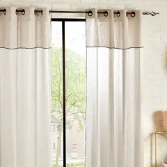 Helle Vorhänge mit schwarzer Umrandung sind an einer Wand befestigt. Ein Fenster mit Blick ins Grüne ist teilweise verdeckt.