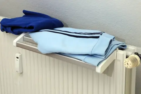 Blaue und hellblaue Kleidung trocknet auf einem Heizkörper. Ein kleiner weißer Kasten mit Display hängt an der Wand.