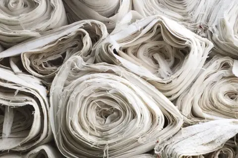 Stapel von weißem, zerknittertem Stoff, der an aufgerollte Papierrollen erinnert. Die Textur ist faserig und unregelmäßig, was auf die Herstellung von Rayonstoff hindeutet, der schädlich für die Umwelt sein kann.