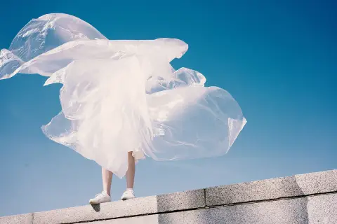 Eine Person steht auf einer Mauer, umgeben von wehendem, weißem Material, das an Plastik erinnert. Polyester schädlich für die Umwelt.