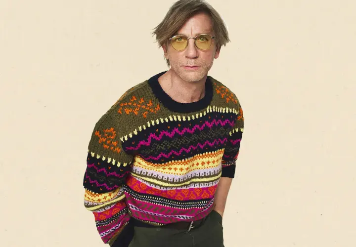 Mann mit gelber Brille trägt bunten wollpullover. Er fragt sich, wie er diesen am besten waschen kann, um die Farben zu erhalten.