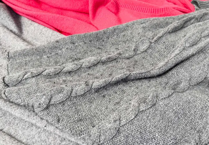 Grauer Strickpullover mit Zopfmuster und ein pinker Stoff. Kleine Knötchen zeigen, wie man Pilling vermeiden kann.