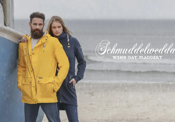 Mann und Frau am Strand im Regen, er trägt eine gelbe Schmuddelwedda Jacke, sie eine blaue. Perfekt für jedes Wetter!