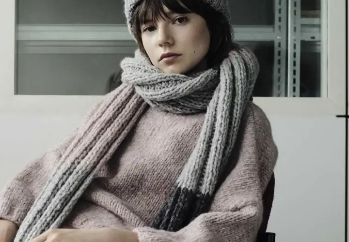 Junges Mädchen mit einem gestrickten Schal aus Wolle und passender Mütze. Der dicke, graue Schal ist um den Hals geschlungen und reicht bis zum rosafarbenen Pullover.