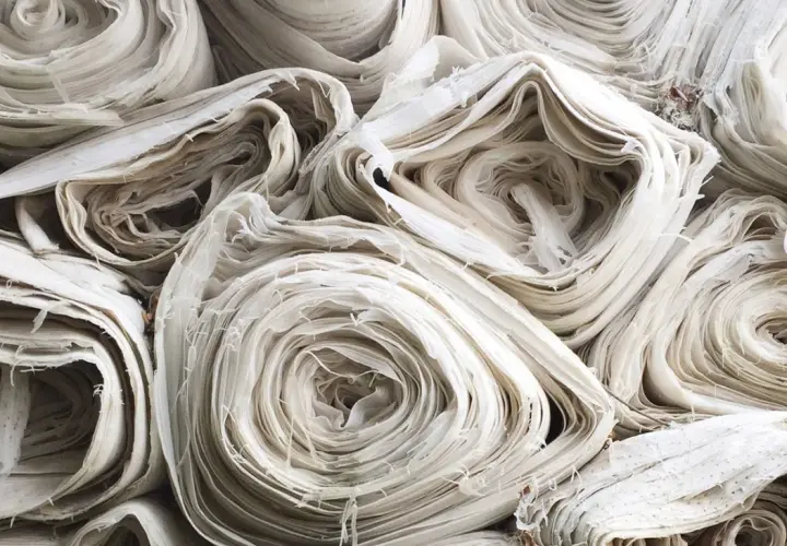 Stapel von weißem, zerknittertem Stoff, der an aufgerollte Papierrollen erinnert. Die Textur ist faserig und unregelmäßig, was auf die Herstellung von Rayonstoff hindeutet, der schädlich für die Umwelt sein kann.