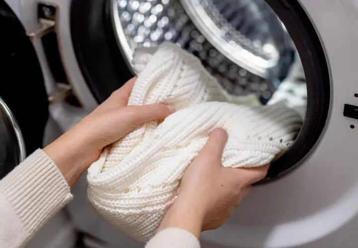 Hände legen einen weißen Strickpullover in die Waschmaschine, um eingelaufene Kleidung zu retten.