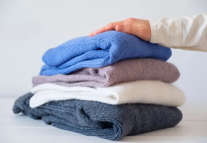 Ein Stapel gestrickter Pullover in verschiedenen Farben, von dunkelblau bis weiß. Eine Hand streicht sanft über den obersten Pullover, als ob er per Hand gewaschen wurde.