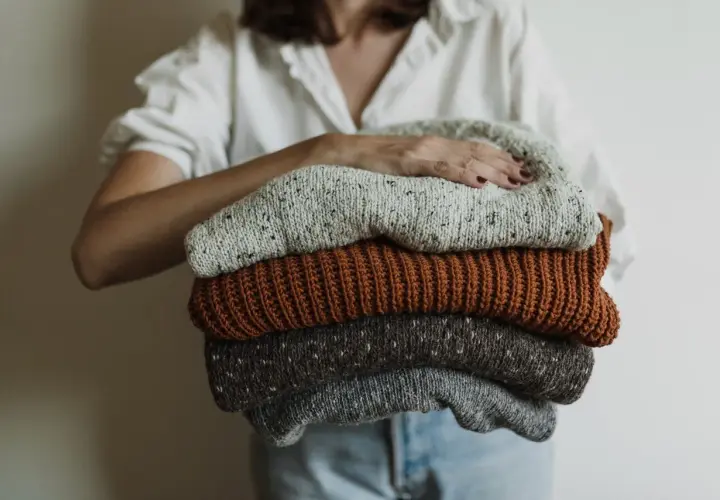 Eine Person hält einen Stapel gestrickter Pullover. Die Pullover sind in Grau-, Braun- und Beigetönen gehalten. Sie sind bereit, um **Wolle bei 40 Grad waschen** zu können.