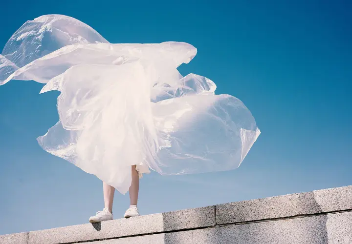 Eine Person steht auf einer Mauer, umgeben von wehendem, weißem Material, das an Plastik erinnert. Polyester schädlich für die Umwelt.