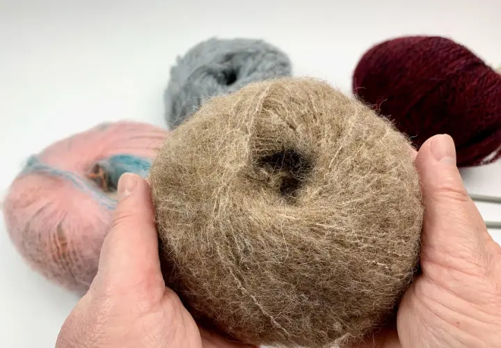 Hände halten ein Knäuel beiger Wolle, bereit, einen Schal zu stricken. Weitere Wollknäuel in Rosa, Grau und Bordeauxrot liegen im Hintergrund.