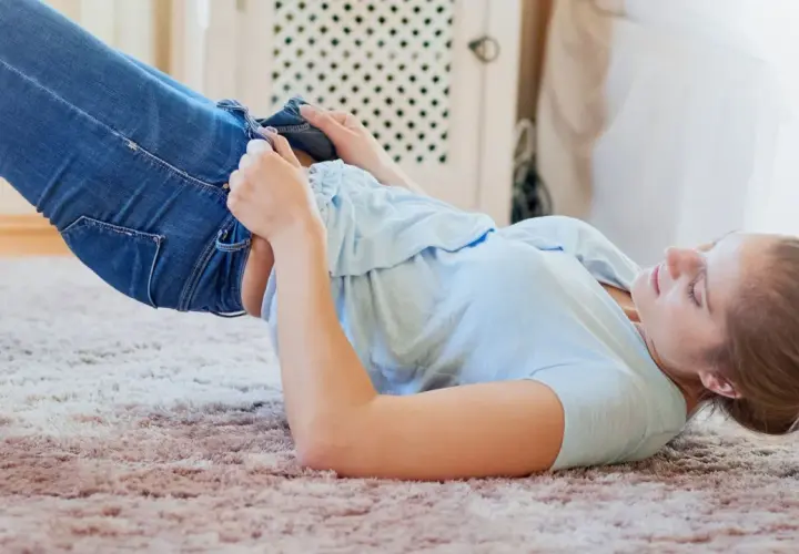 Frau kämpft mit ihrer Jeans, die eingelaufen ist. Was tun? Sie liegt auf einem Teppich und versucht, die Hose anzuziehen.