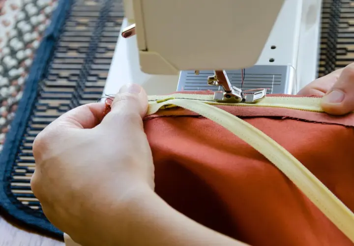 Hände nähen einen Reißverschluss mit einer Nähmaschine an ein orangefarbenes Kleidungsstück. Reißverschluss reparieren leicht gemacht.
