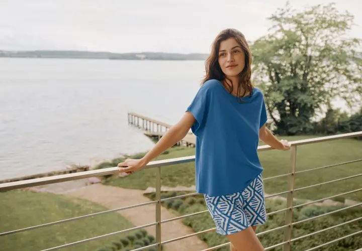 Frau auf Balkon mit Blick aufs Wasser, trägt blaues Oberteil und gemusterte Shorts aus Modal Stoff. Waschen ist einfach.
