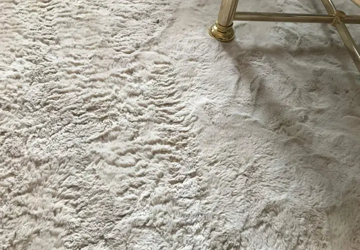 Ein heller, flauschiger Kunstfell Teppich ist leicht verfilzt. Ein goldener Möbelfuß steht darauf.