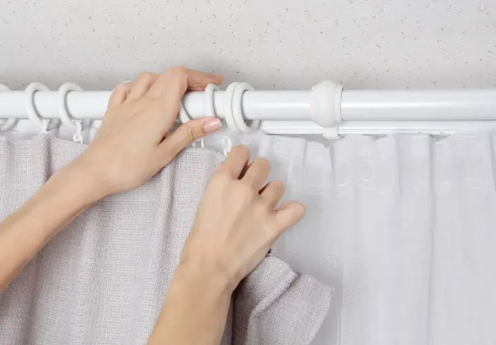 Hände befestigen zwei Gardinen auf einer Stange. Eine Gardine ist grau, die andere weiß und transparent.