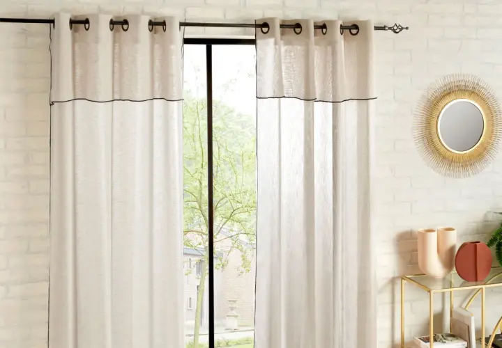 Helle Vorhänge mit schwarzer Umrandung sind an einer Wand befestigt. Ein Fenster mit Blick ins Grüne ist teilweise verdeckt.