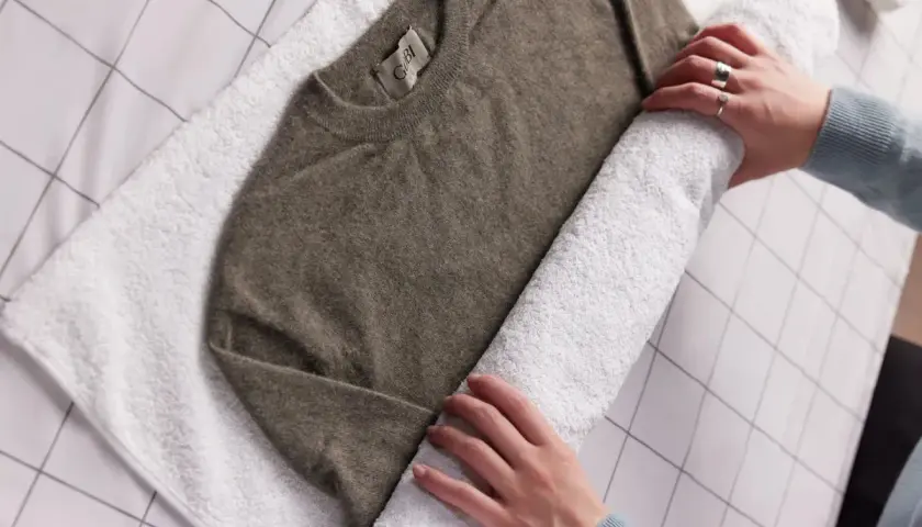 Ein brauner Kaschmirpullover wird auf einem weißen Handtuch gerollt, um ihn nach dem Waschen mit Essig zu trocknen.