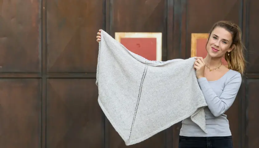 Junge Frau hält ein hellgraues, gestricktes Dreieckstuch hoch.