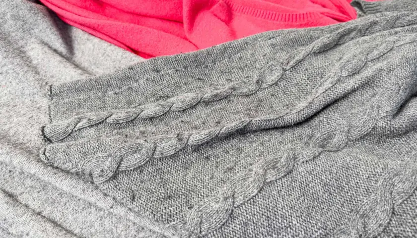 Grauer Strickpullover mit Zopfmuster und ein pinker Stoff. Kleine Knötchen zeigen, wie man Pilling vermeiden kann.