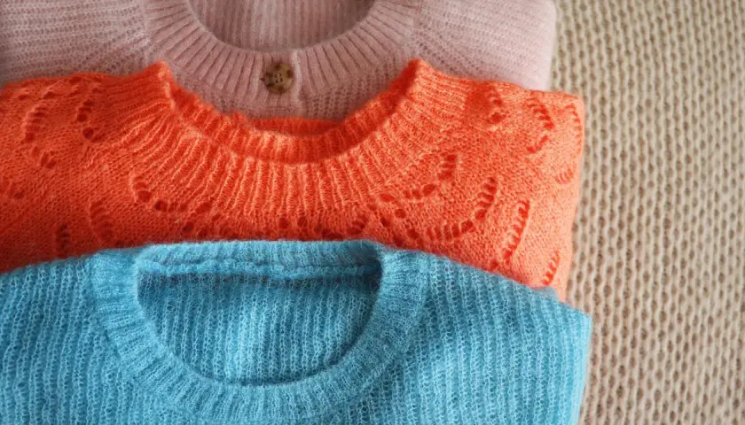 Drei gestapelte Pullover in Rosa, Orange und Blau. Die feine Lammwolle ist bereit für das Waschen, um ihre Weichheit zu erhalten.
