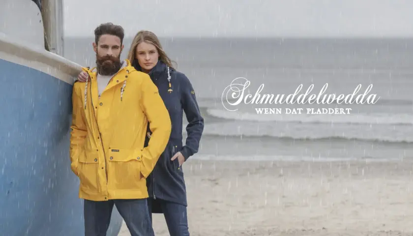 Mann und Frau am Strand im Regen, er trägt eine gelbe Schmuddelwedda Jacke, sie eine blaue. Perfekt für jedes Wetter!