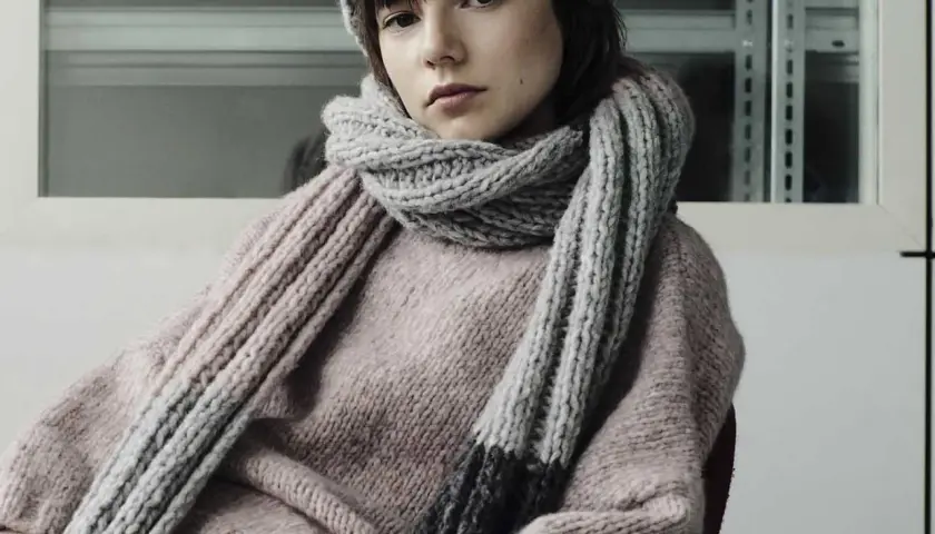Junges Mädchen mit einem gestrickten Schal aus Wolle und passender Mütze. Der dicke, graue Schal ist um den Hals geschlungen und reicht bis zum rosafarbenen Pullover.