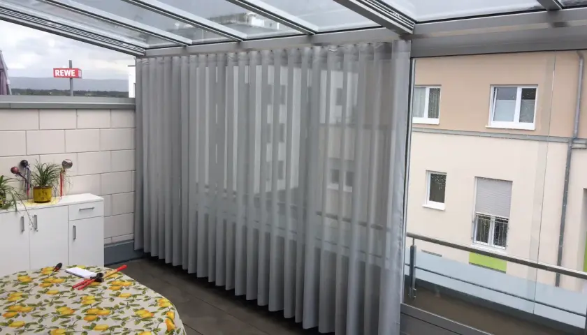 Gardinen im Wintergarten befestigen: Helle Stoffbahnen hängen von einer Schiene unter dem Glasdach.